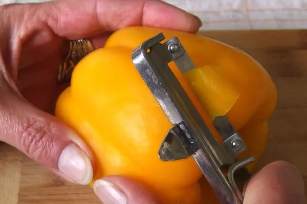 Eine Hand schält eine gelbe Paprika mit einem Schäler auf einem Holzbrett in einer hellen Küchenumgebung.