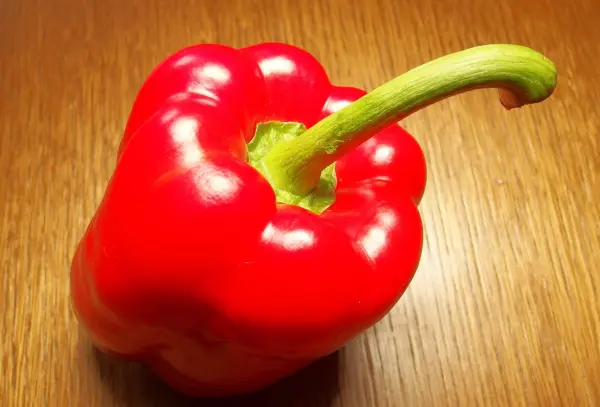 Eine rote Paprikaschote liegt auf einem Holztisch, bereit zum Schälen und Zubereiten für ein Gericht.
