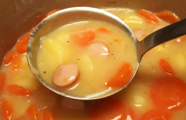 Ein Löffel nimmt eine milchige Suppe mit Karotten und Würstchen auf, die in einem Topf zubereitet wird. Versalzene Speisen können den Genuss mindern.