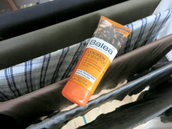 Haarshampoo liegt auf einem Wäscheständer mit Kleidung. Text auf der Flasche: “Balea PROFESSIONAL LOCKEN SHAMPOO. Von Friseuren empfohlen. Lockiges und gewelltes Haar.”