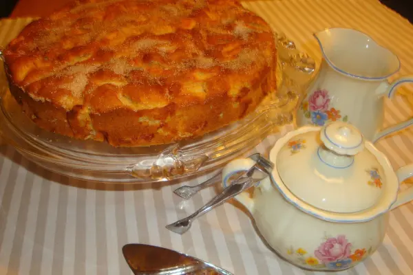 Ein saftiger Apfelkuchen steht in einer Glasplatte, umgeben von einer schönen Teekanne und einer Milchkanne auf einem gestreiften Tisch.