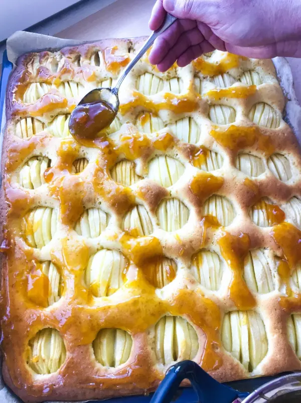 Ein handgehaltener Löffel verteilt Marmelade auf einem frisch gebackenen Blitz-Apfelkuchen in der Auflaufform.