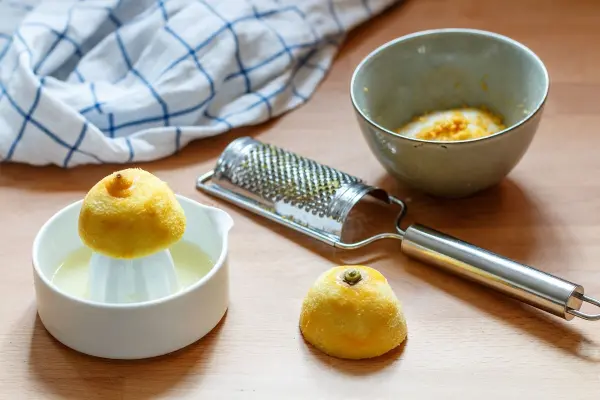 In der Szene stehen frische Zitronen auf einem Holztisch neben einer Schüssel und einer Reibe; sie bereiten ein "Koldskal"-Dessert vor.