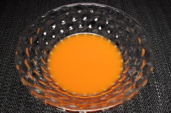 Ein tiefes Schüsselchen mit leuchtend orangefarbigem Saft steht auf einer dunklen Oberfläche und reflektiert sanft das Licht.