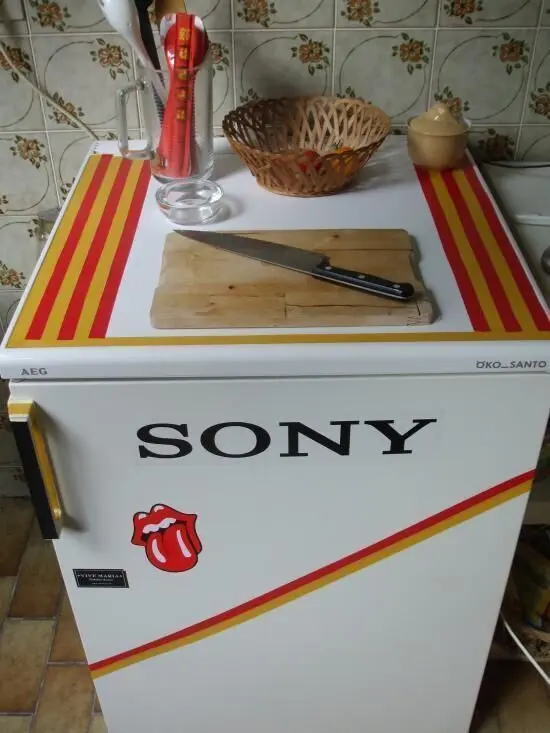 Ein verzierter Kühlschrank hat bunte Streifen und Sticker, steht in einer Küche mit Schneidebrett, Messer und Utensilien darauf. Text: "SONY".