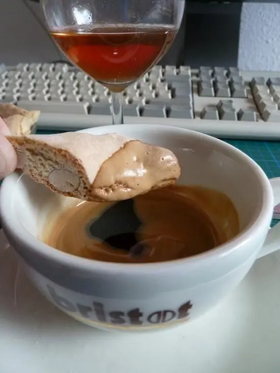 Eine Cantuccini wird in eine Tasse Kaffee getunkt, mit einem Weinglas und einer Computertastatur im Hintergrund. Text auf der Tasse: "bristat".