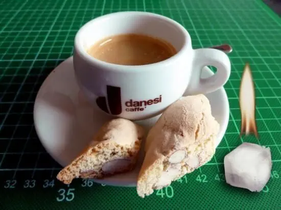 Ein Kaffee in einer weißen Tasse liegt neben zwei Cantuccini, alles auf einem grünen Untergrund mit Maßstab.