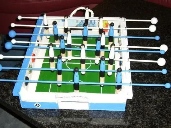 Tischfußballspiel mit Spielfiguren auf grünem Feld; gebaut auf schwarzem Tisch. Holzstangen halten blau-weiße Spielerfiguren.