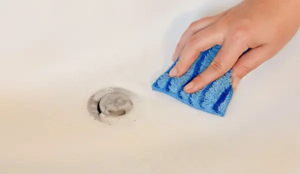 Eine Hand mit einem blauen Schwamm reinigt eine Badewanne neben einem Abfluss, um das Essigreiniger-Ergebnis zu verbessern.