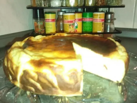Käsekuchen ohne Boden sitzt auf einem Glasteller in der Küche, mit einer herausgeschnittenen Scheibe. Im Hintergrund Gewürze.