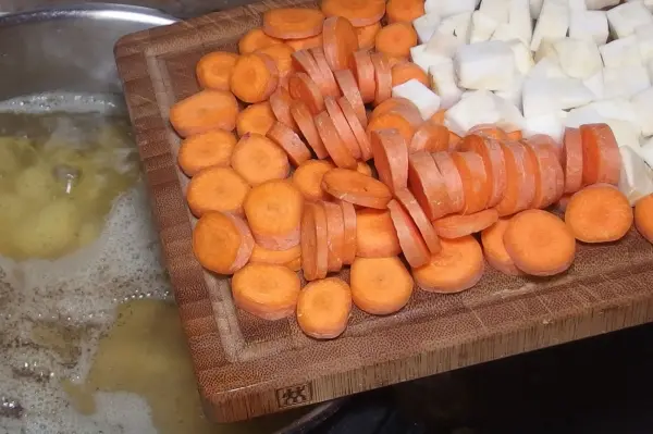Auf einem Holzbrett liegen geschnittene Möhren und Kohlrabi, während im Hintergrund eine Kartoffelsuppe kocht.