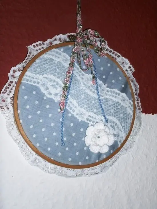 Ein runder, bestickter Stickrahmen mit blauer, gepunkteter Stoffunterlage hängt an einer Wand und zeigt eine filigrane Spitzenborte.