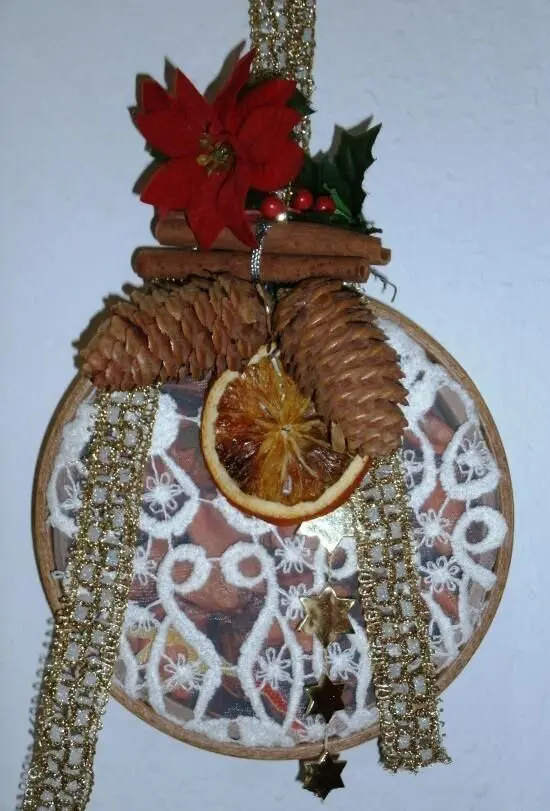 Stickrahmen mit getrockneter Orangenscheibe und Zapfen dekoriert, hängt mit goldenen Bändern und Spitzenmuster.