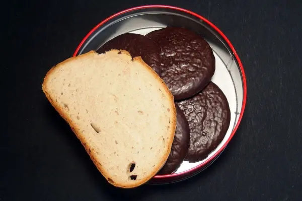 Ein Stück Brot liegt neben einer Auswahl an Plätzchen in einer roten Dose auf schwarzem Hintergrund.