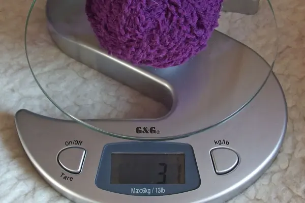 Ein lila Wollknäuel liegt auf einer digitalen Waage, die 37 Gramm anzeigt, auf einem hellen, weichen Untergrund.