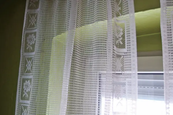 Weiße Gardinen hängen vor einem Fenster, leicht im Licht wehend, umgeben von einem hellgrünen Raum.