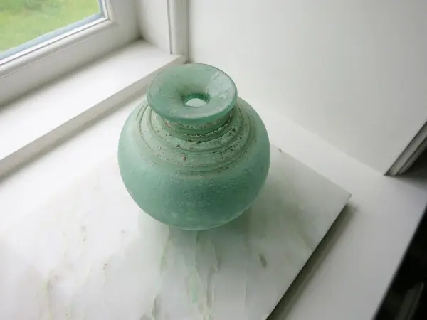Eine grünliche Glasblumen vase steht auf einem weißen Tisch in der Nähe eines Fensters, lichtdurchflutet und sauber.