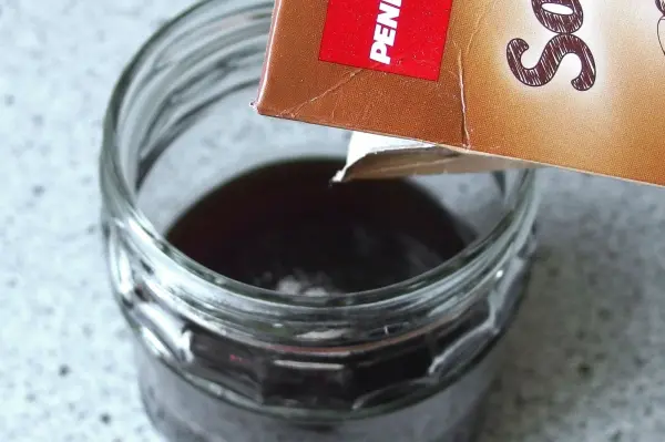 Ein Glas mit dunkler Flüssigkeit wird von einem Karton mit der Aufschrift "Sahne" übergossen. Die Küchensituation deutet auf Kochaktivität hin.