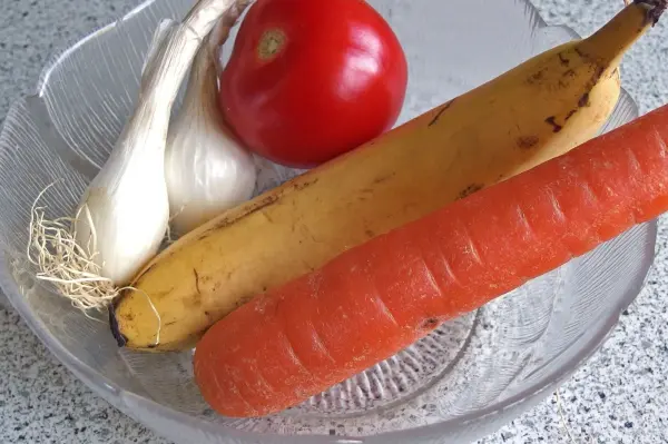 Ein Teller enthält eine Tomate, eine Banane, eine Möhre und eine Knoblauchzehe, die frisch und bunt angeordnet sind.