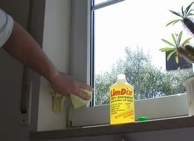 Eine Person reinigt weiße Fensterrahmen mit einem Tuch. Daneben steht eine Flasche "limDor" Grillreiniger auf der Fensterbank.