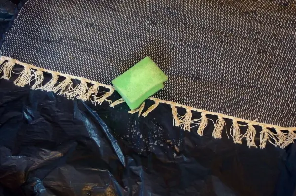 Ein Teppich liegt auf einem schwarzen Untergrund, während ein grüner Schwamm über die Teppichfransen gereinigt wird.