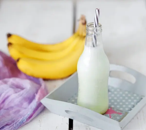 Ein Buttermilch-Bananen-Shake steht in einer Flasche auf einem Tablett, während dahinter frische Bananen liegen.