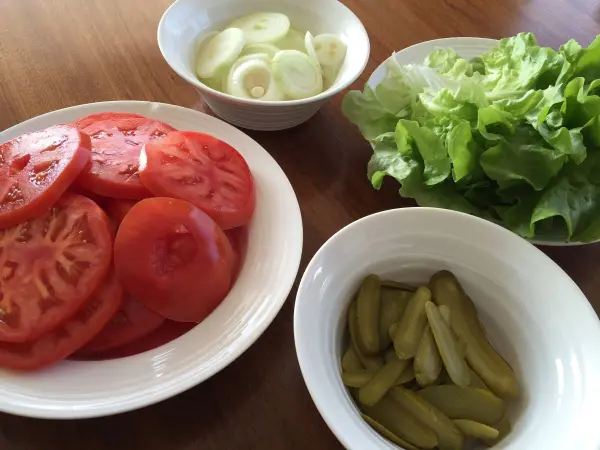 Frische Zutaten wie Tomatenscheiben, Salat, Gurken und Zwiebeln liegen in Schüsseln bereit, um einen Original American Cheeseburger zuzubereiten.