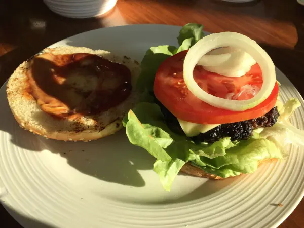 Ein Cheeseburger liegt auf einem Teller, während die obere Brötchenhälfte und die beladenen Zutaten wie Salat und Tomate bereit sind.