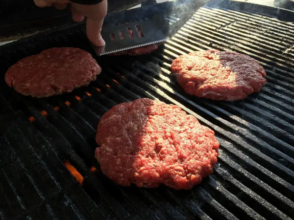 Drei rohe Hamburger-Patties werden auf einem Grill gewendet, um einen Original American Cheeseburger zuzubereiten.