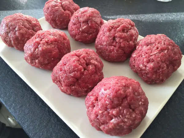 Rohe Hackfleischbällchen liegen auf einem weißen Teller, platziert auf einer dunklen Oberfläche, bereit zum Zubereiten.
