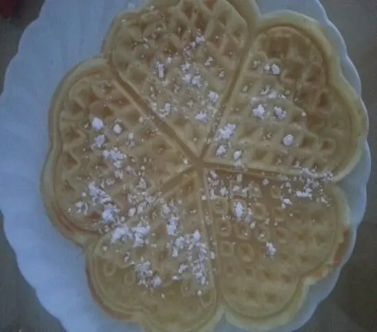 Herzförmige Waffeln mit Puderzucker sind auf einem weißen Teller angerichtet und liegen auf einer neutralen Oberfläche.