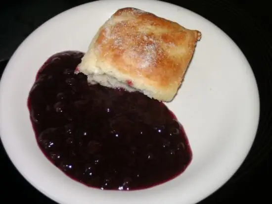 Eine Quarkbuchtel liegt auf einem weißen Teller, begleitet von lila fruchtiger Sauce, die großzügig daneben ausgegossen ist.