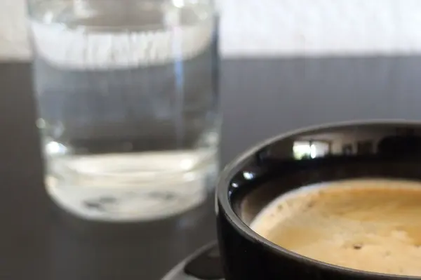 Eine schwarze Tasse mit Kaffee steht auf einem Tisch, während im Hintergrund ein Glas Mineralwasser sichtbar ist.