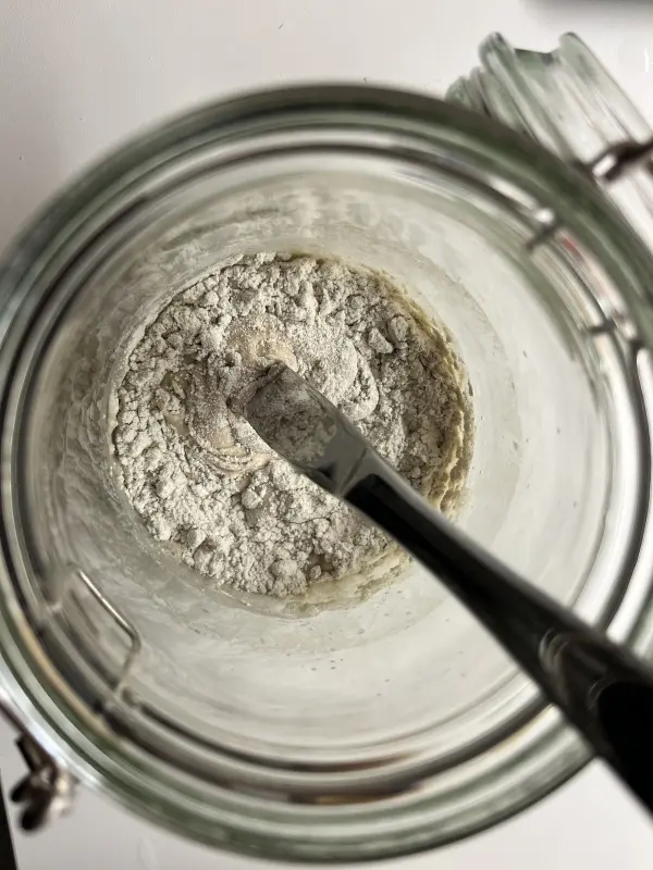 Ein Glas enthält Sauerteig mit Mehl und einer Mischung, während ein Löffel umrührt. Helle Küchenumgebung.