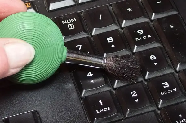 Ein kleiner, grüner Blasebalg mit Pinsel reinigt ein Keyboard, während eine Hand das Gerät führt.