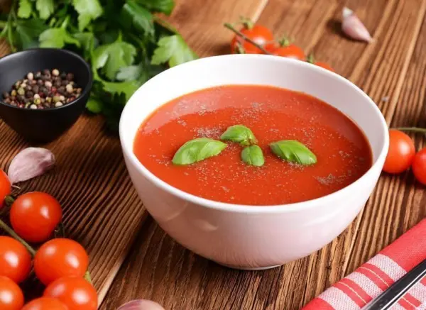 Tomatensuppe in weißer Schale, garniert mit Basilikum, umgeben von Tomaten, Pfeffer und Kräutern auf Holztisch.