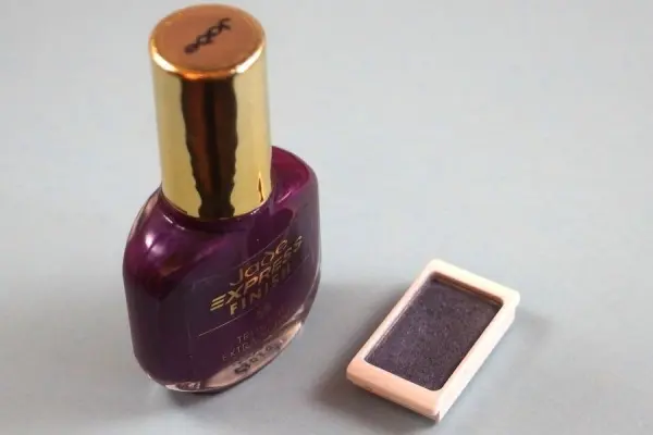 Ein Nagellack in kräftigem Lila steht neben einem alten Lidschatten in einem kleinen, rechteckigen Behälter auf einem neutralen Untergrund.