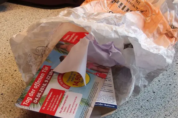 Eine zerrissene Bäckertüte liegt in einer Schüssel, während darin auch bunte Werbeflyer zu sehen sind, die Abfall zeigen.