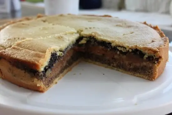 Der Gedeckte Nusskuchen zeigt ein aufgeschnittenes Stück, enthüllt dreierlei Füllungen. Er steht auf einer weißen Platte.