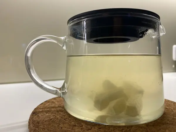 Ein transparenter Becher enthält eine helle Flüssigkeit mit Ingwerstücken, die auf einem Untersetzer steht.