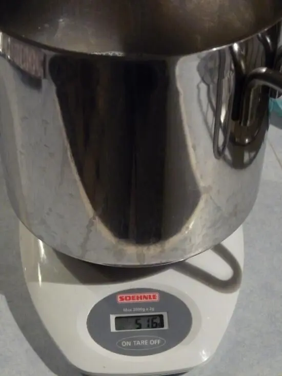 Eine Küchenwaage wiegt einen großen, glänzenden Metalltopf und zeigt ein Gewicht von 5 kg an, auf einer Arbeitsfläche.