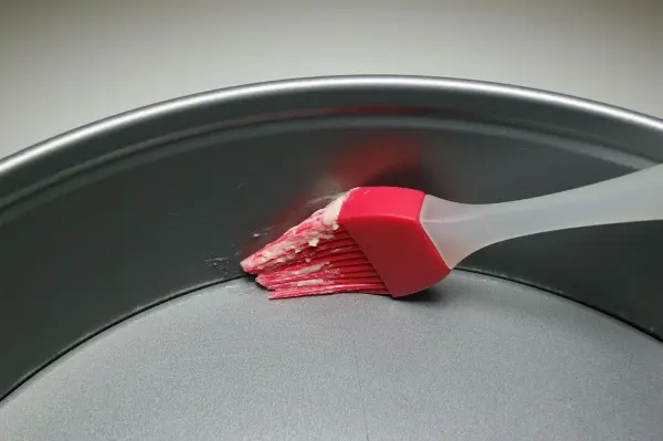 Ein Pinsel mit rotem Silikon fettiert gleichmäßig die Innenfläche einer runden Backform aus Metall, vor einem neutralen Hintergrund.