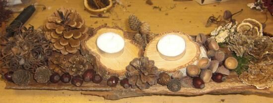Zwei Kerzen stehen auf einer rustikalen Holzplatte, umgeben von Weihnachtsdeko aus Naturmaterialien wie Zapfen und Nüssen.