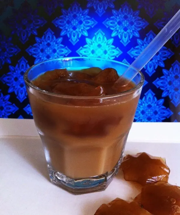 Ein Glas Eiskaffee steht auf einem hellen Untergrund, gefüllt mit gefrorenem Kaffee und einem Strohhalm, vor einem bunten Hintergrund.
