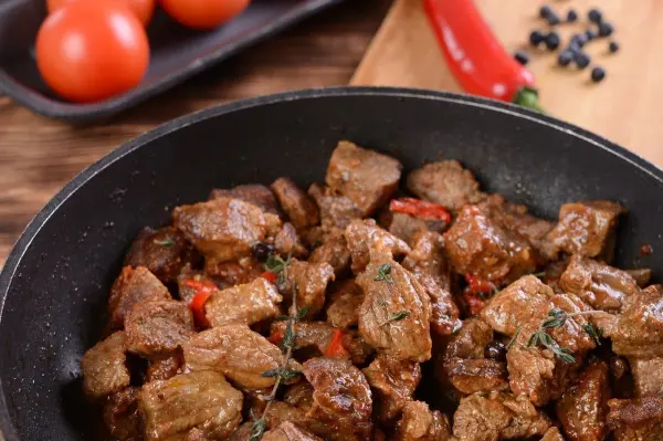 Ein Gulasch aus dem Backofen kocht in einer Pfanne, umgeben von frischen Tomaten und Gewürzen auf einem Holzbrett.