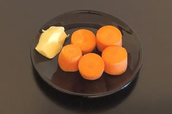 Fünf leuchtend orangefarbene Möhrenstücke liegen auf einem schwarzen Teller, neben einem Stück Fett.