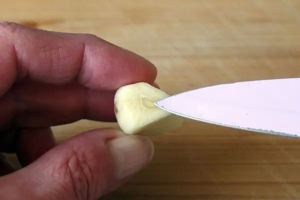 Eine Hand hält eine Knoblauchzehe, während ein Messer an die Zehe angesetzt wird, um den unangenehmen Knoblauchgeruch zu reduzieren.