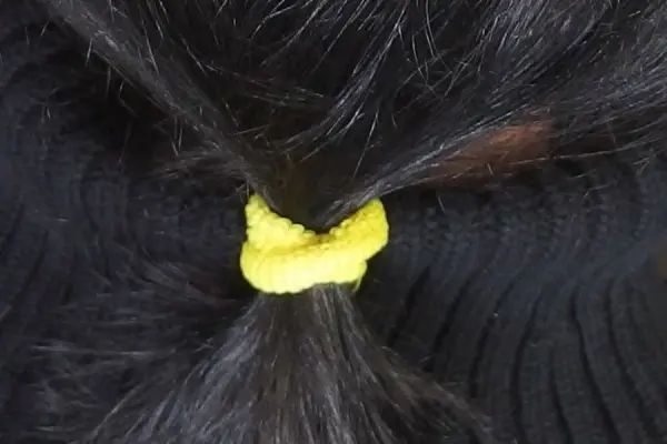 Ein gelbes Haargummi ohne Metallstück hält lange, schwarze Haare zu einem Pferdeschwanz gebunden, vor einem schwarzen Hintergrund.
