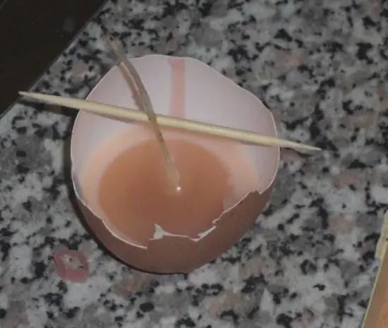 Eine Eierkerze befindet sich in einer Eierschale, während Wachs darin gegossen wird. Ein Holzstäbchen hält den Docht in Position.
