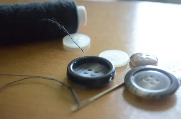 Ein schwarzer Knopf mit vier Knopflöchern liegt auf einem Tisch, während ein Faden und eine Nadel bereit zum Annähen sind.